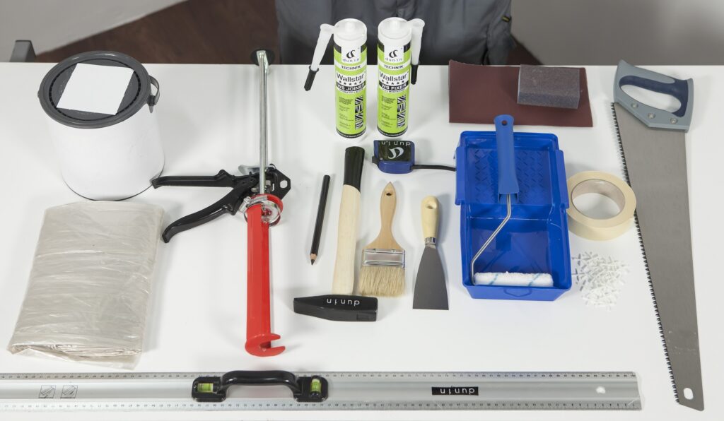 Jak montować listwy z poliuretanu? Obraz przedstawia stół, na którym przygotowano niezbędne rzeczy do zamontowania listwy. Kleje, fugi, tacka i wałek malarski.
